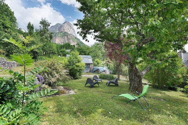 A grassy outdoor area with a tree, a small building, and three chairs.
