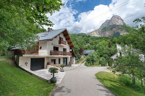 A two-story house with a balcony, a garage, and a driveway leading to a mountain.