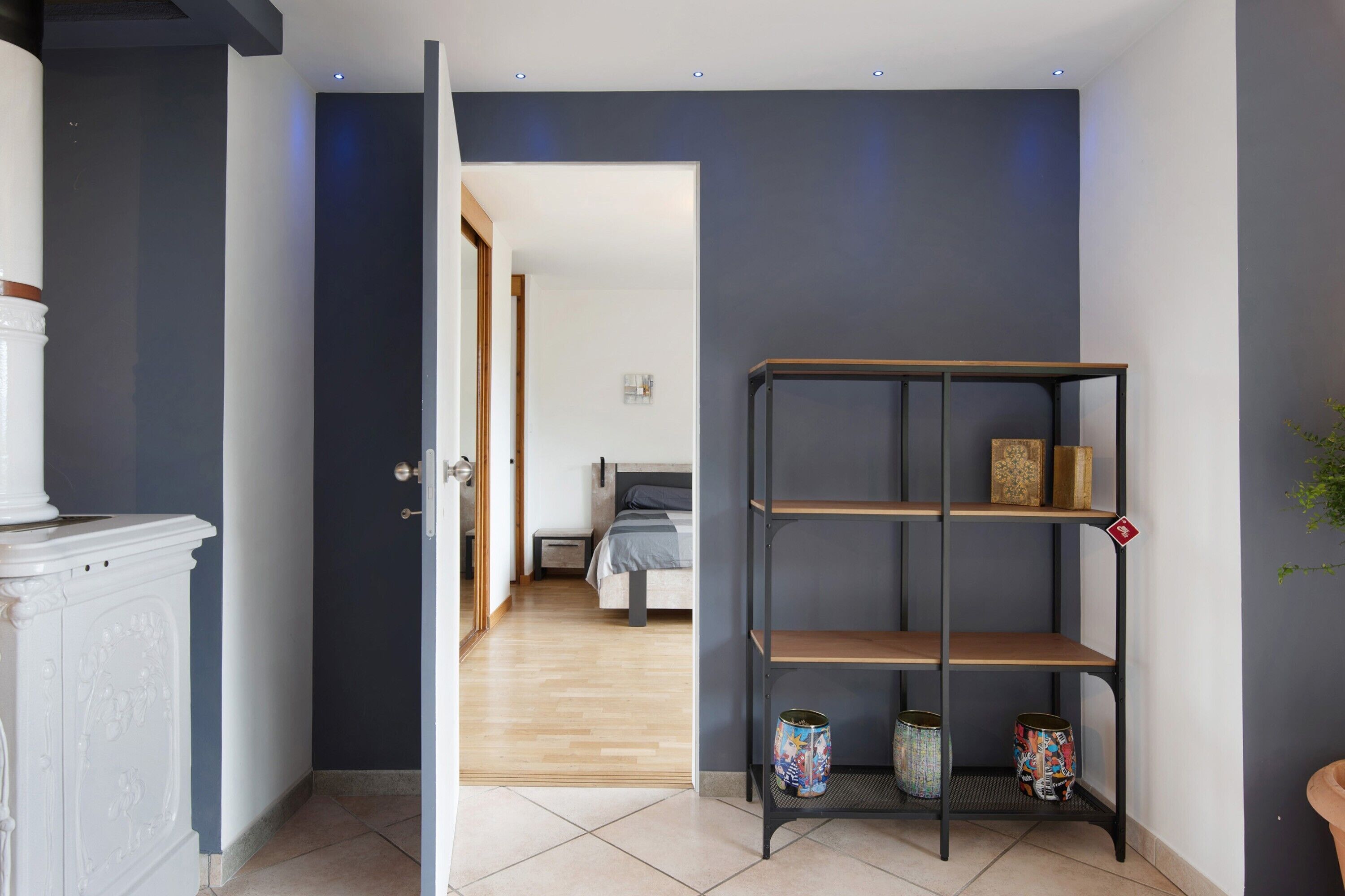 A hallway with a white cabinet, a black metal shelf with decorative items, and a bedroom in the background.