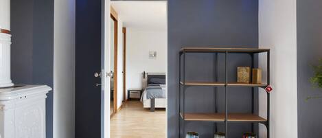 A hallway with a white cabinet, a black metal shelf with decorative items, and a bedroom in the background.