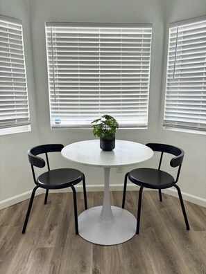 Quarto com mesa redonda, duas cadeiras, planta em vaso e janelas amplas com persianas.