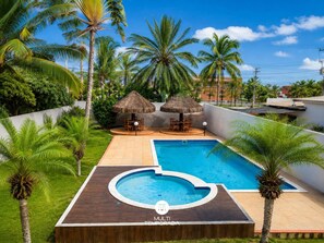 Ein Pool mit Holzterrasse, umgeben von Palmen und einer Sitzgelegenheit mit einem Reetdach.
