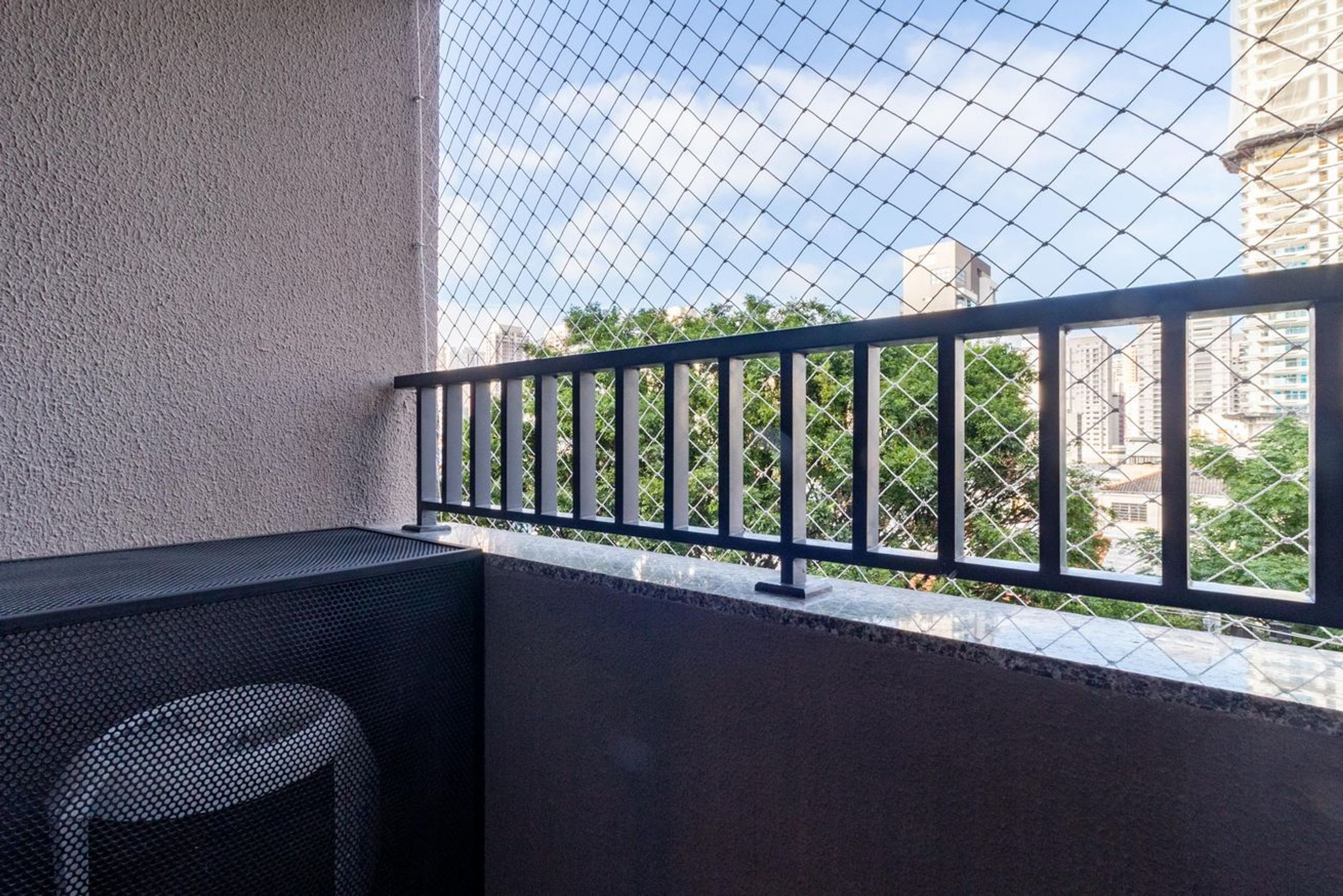 Ein Balkon mit einem Drahtzaun, einem kleinen Tisch und Blick auf Gebäude und Bäume.
