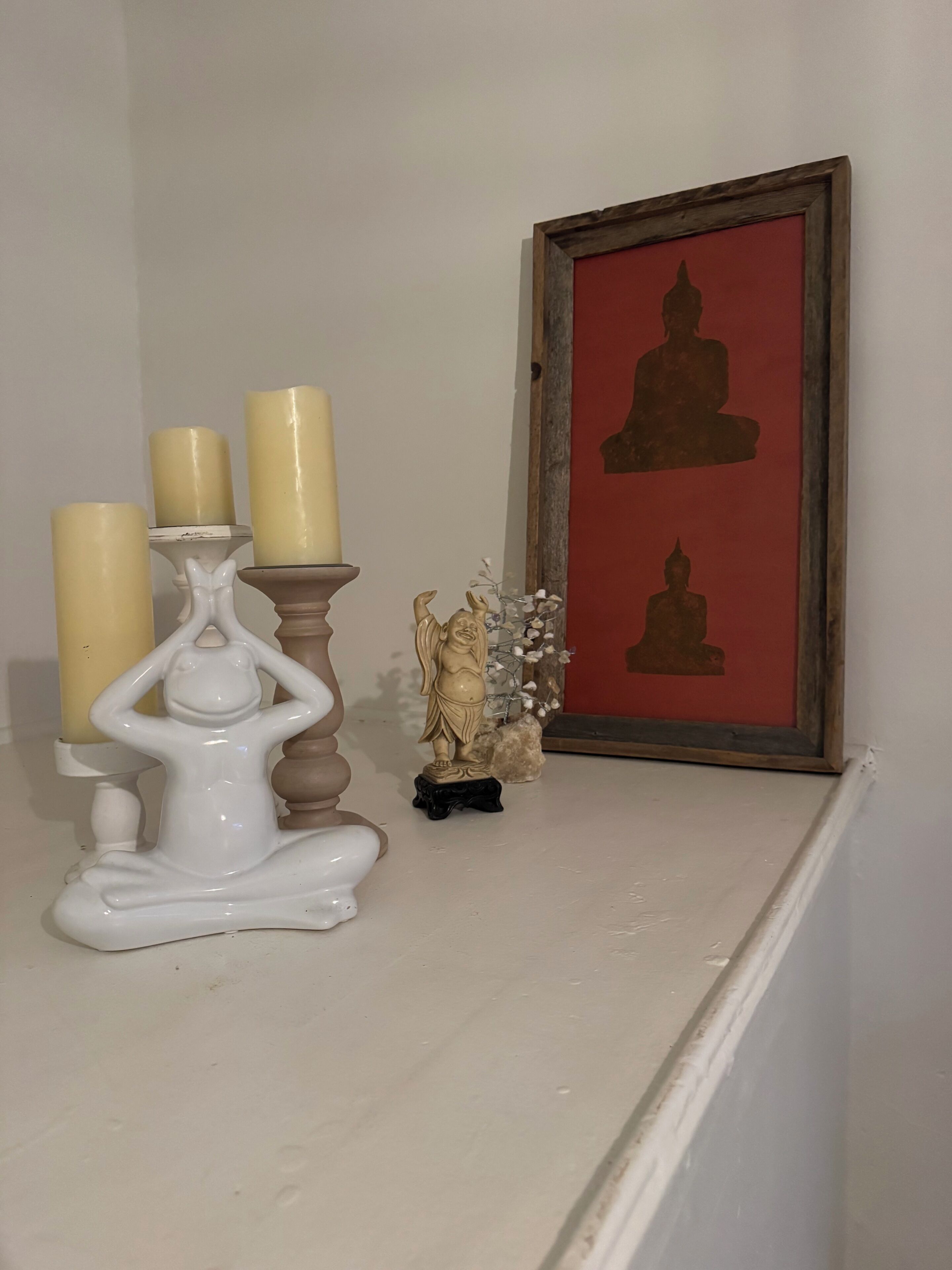 Uno scaffale con una figura in ceramica bianca, tre candele, una piccola statua e un quadro incorniciato.
