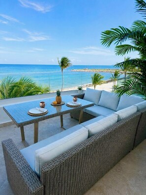 A wicker sofa with a glass table and chairs, a pineapple, and a pitcher, overlooking a beach with palm trees and the ocean. - OCEANFRONT Punta Palmera Paradise ~ 2 BR apt. in Cap Cana w/Private Beach Access (Punta Cana)