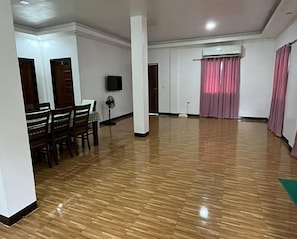 A spacious room with a wooden floor, a dining area with chairs and a table, a standing fan, and an air conditioning unit.