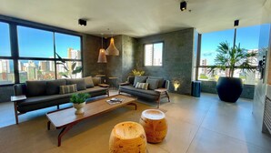 A modern living room with a sofa set, coffee table, and two wooden stools. Large windows offer a city view. - Condominio Jsmart, Localizacao Ideal (Fortaleza)