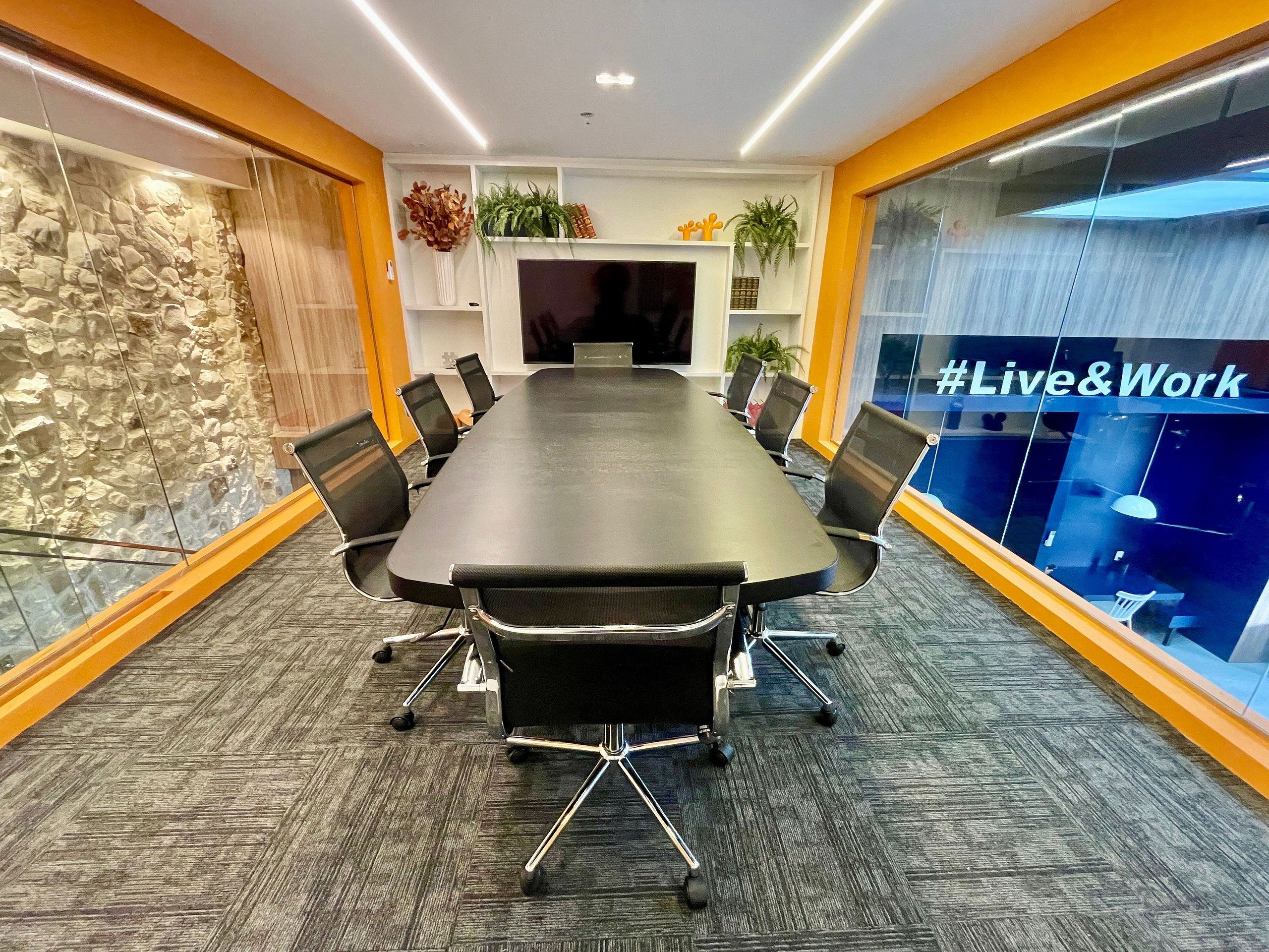 A conference room with a long table, chairs, a TV screen, and a glass wall with the text '#Live&Work'.
