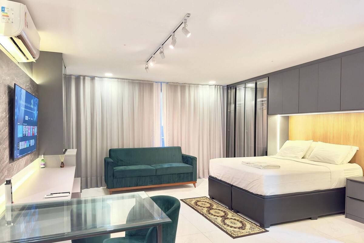 Quarto de hotel moderno com cama, sofá verde, televisão e mesa de vidro.
