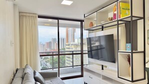 A modern living room with a large TV, a sofa, a bookshelf, and a city view through a large window. - Condominio Jsmart, Localizacao Ideal (Fortaleza)