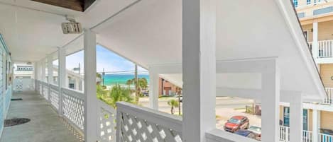 A covered balcony with a view of the beach and ocean, white railings, and a clear sky.