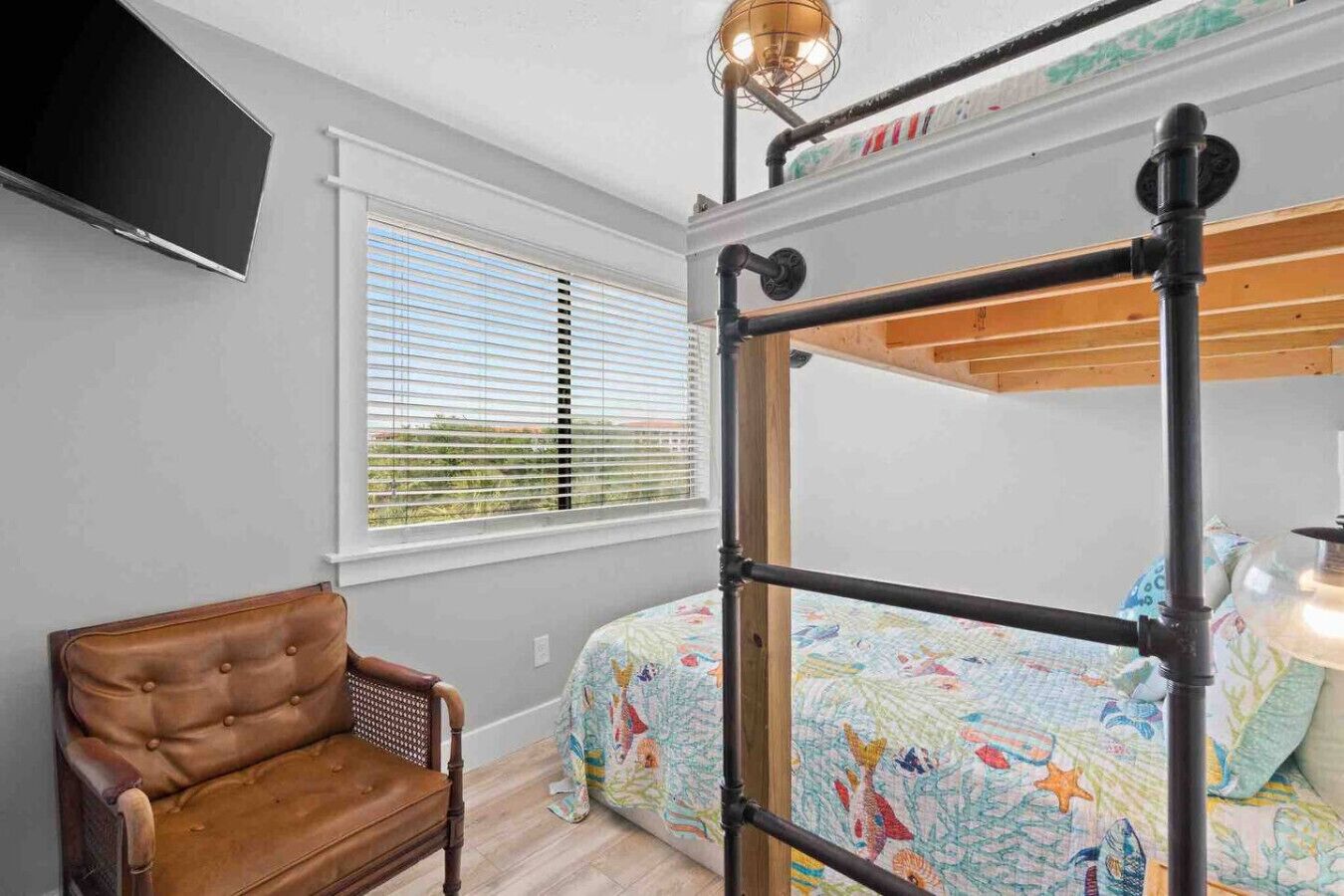 A bedroom with a bunk bed, a leather chair, a TV mounted on the wall, and a window with blinds.
