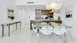 A modern kitchen with a glass dining table and white chairs, a chandelier, and a bench. - Deluxe Brickell Suite w/ pool, gym, & city views! (Miami)