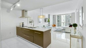 A modern kitchen with a central island, a dining area with a glass table, and a large window offering a city view. - Deluxe Brickell Suite w/ pool, gym, & city views! (Miami)