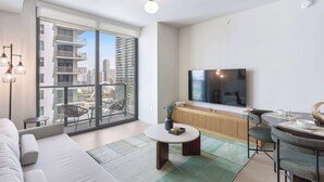 Ein modernes Wohnzimmer mit einer Couch, einem runden Couchtisch, einem Essbereich mit Tisch und Stühlen, einem Flachbildfernseher und einem großen Fenster mit Blick auf die Stadt.