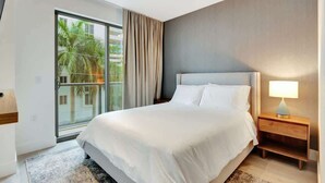 A hotel room with a large bed, a nightstand with a lamp, a sliding glass door to a balcony, and a view of palm trees. - Modern Studio w/ Balcony Near Brickell & Downtown (Miami)