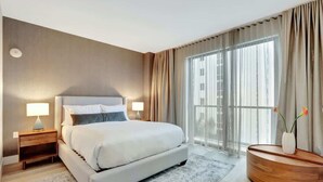 Una habitación de hotel moderna con una cama grande, mesitas de noche, una consola redonda de madera y un amplio ventanal con cortinas.
