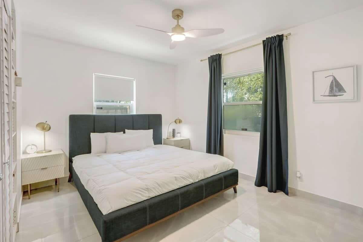 Una camera da letto moderna con un letto, comodini, una ventola a soffitto, una finestra con tende e un quadro appeso al muro.