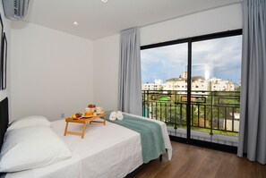 A bedroom with a bed, a small table with a tray, a green blanket, and a view of buildings through a large window. - Residencial Germany (Florianópolis)
