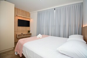 A hotel room with a bed, a flat-screen TV mounted on the wall, a wooden cabinet, and a large window with curtains. - Residencial Germany (Florianópolis)