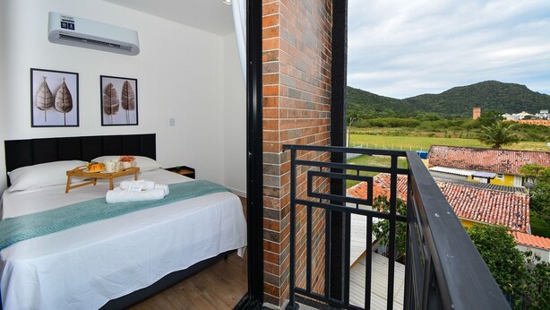 Quarto com cama, mesa de cabeceira com bandeja, parede branca, ar condicionado e varanda com vista para uma paisagem montanhosa.