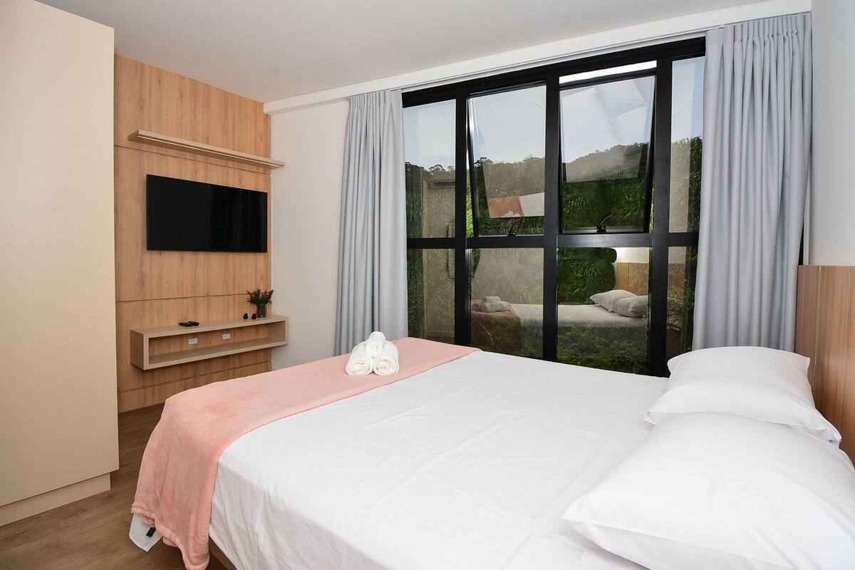 A hotel room with a bed, a flat-screen TV mounted on the wall, a window with curtains, and a small shelf with a plant.