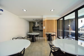 A modern dining area with round tables and black chairs, a kitchen in the background, and large windows with a view of the outside. - Residencial Germany (Florianópolis)