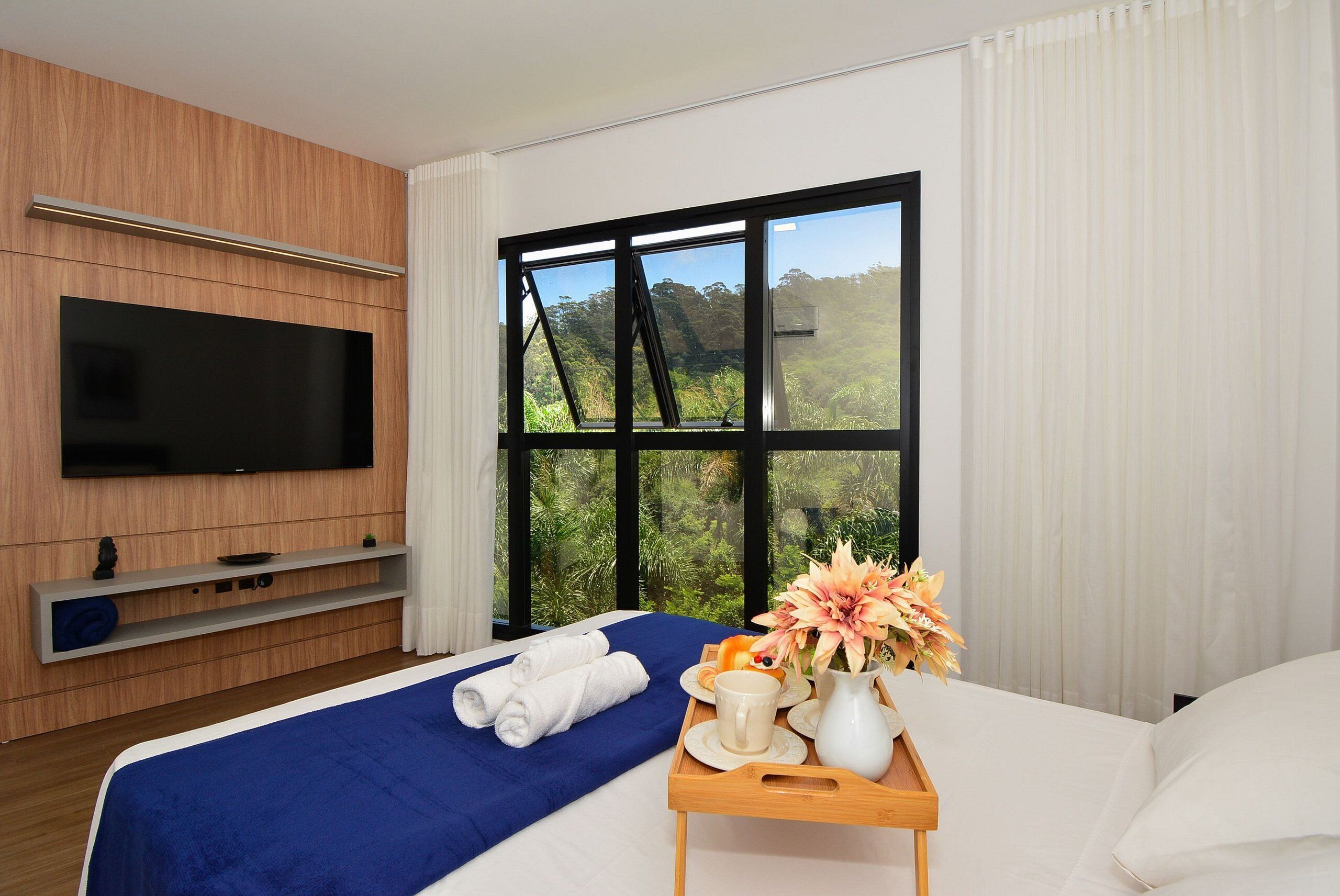 A hotel room with a bed, a TV, a window with a view of greenery, a tray with a vase of flowers, and a breakfast tray.