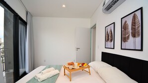 Quarto de hotel com cama, mesa de cabeceira com bandeja de café da manhã, janela com cortinas e duas obras de arte com folhas emolduradas na parede.