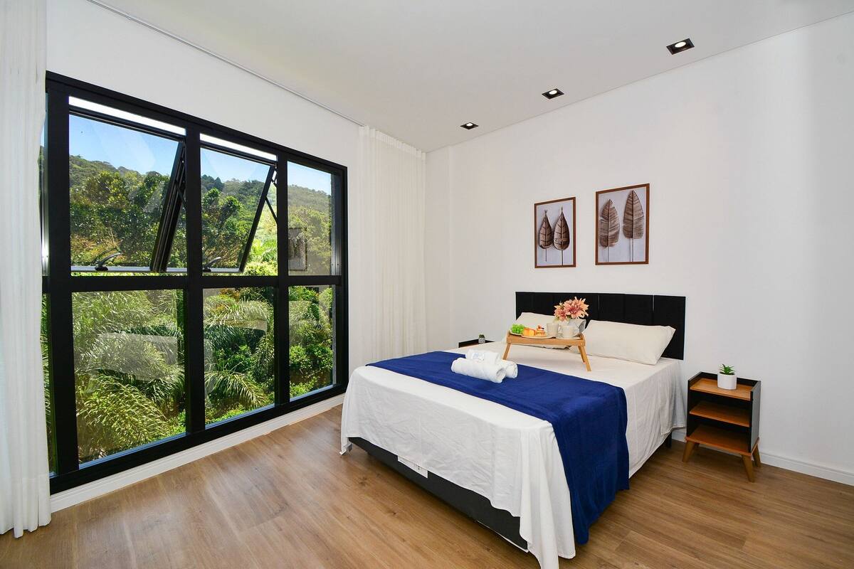 A bedroom with a large window, a bed with a blue bedspread, white bedding, and two framed botanical prints on the wall.