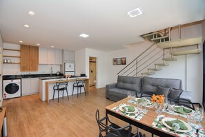 A modern kitchen with a central island, a dining area with a table set for four, and a staircase leading to an upper level. - Residencial Germany (Florianópolis)