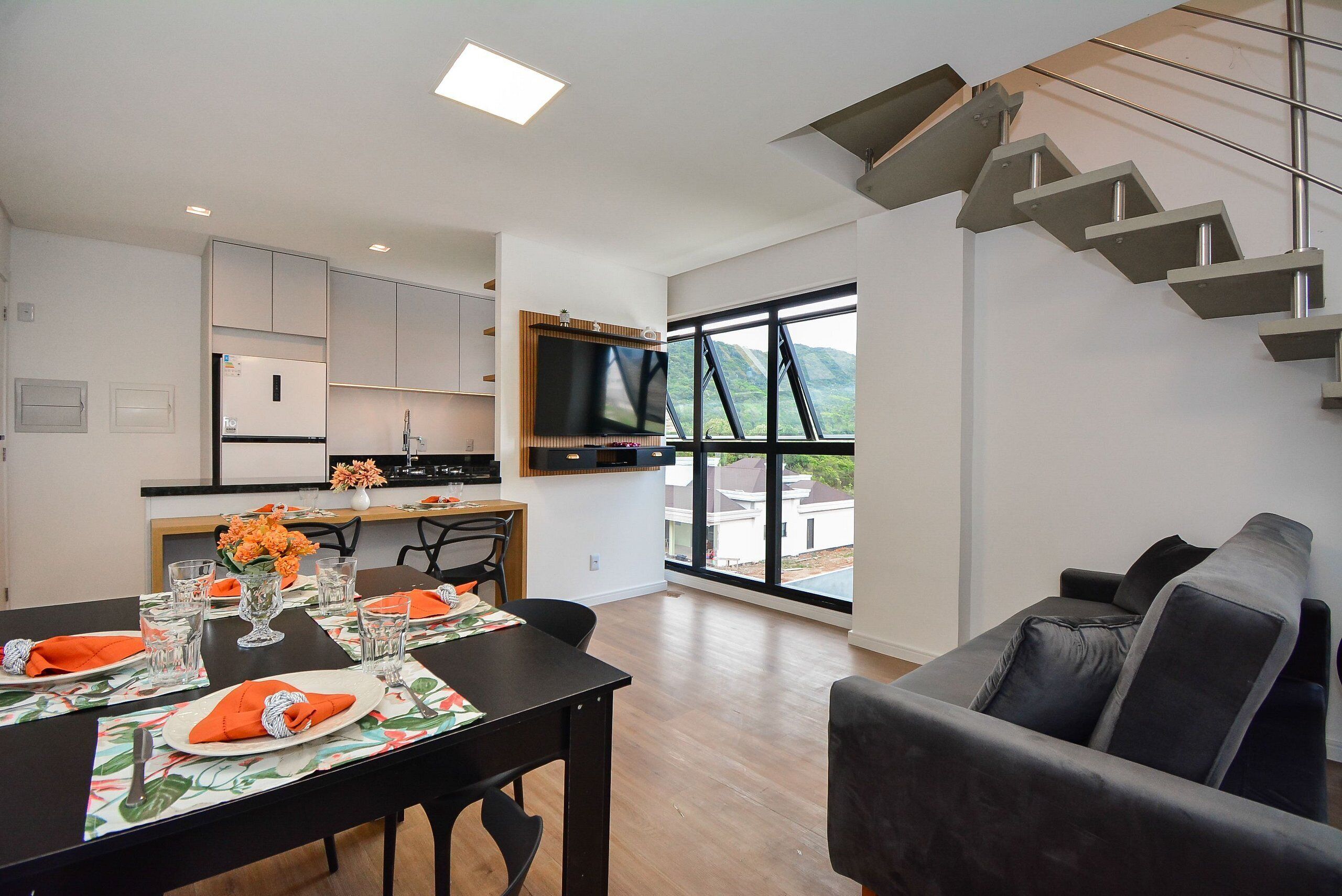 A modern kitchen with a built-in TV, a dining area with a table set for two, and a staircase leading to an outdoor balcony.
