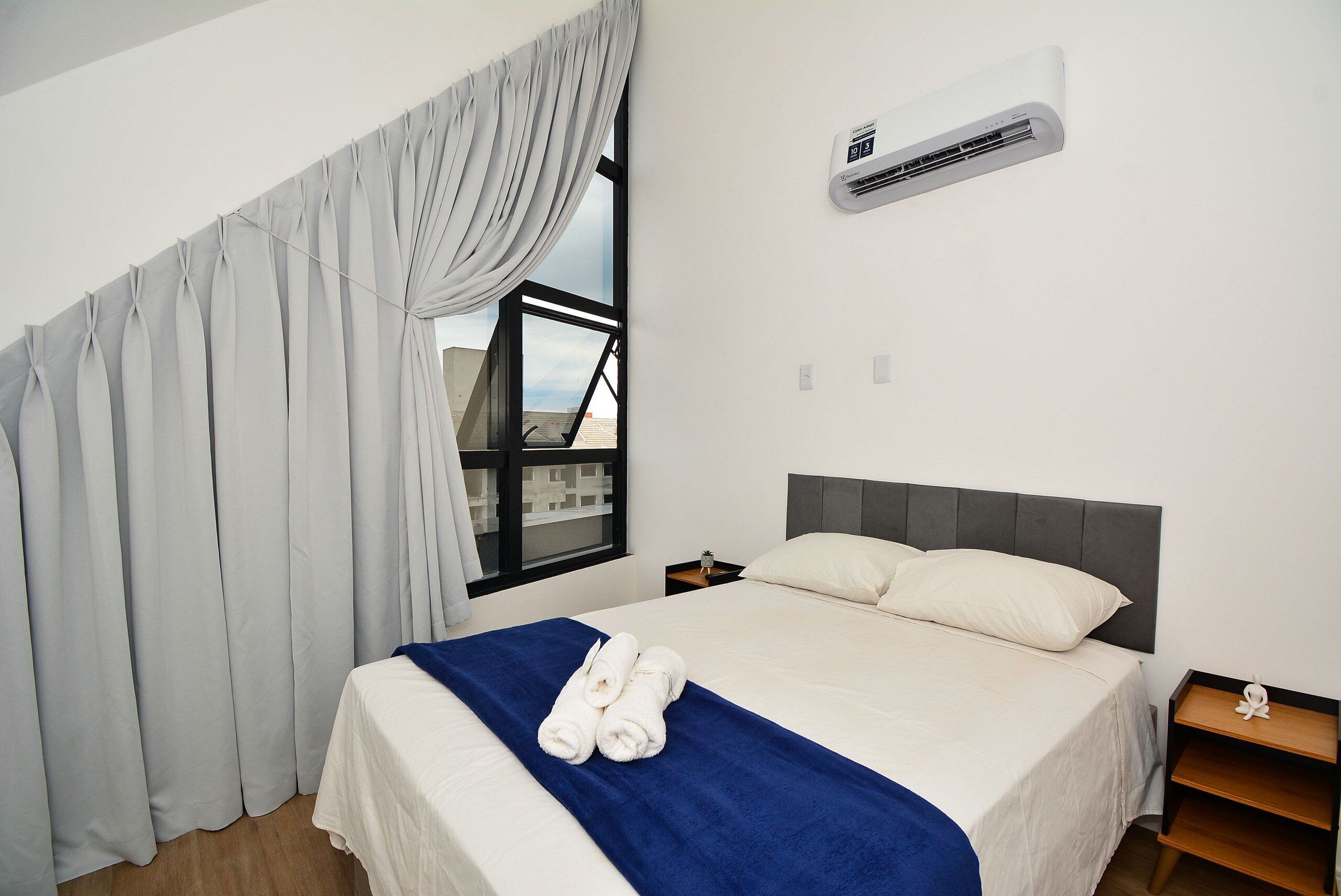 Una habitación de hotel con cama, ventana con cortinas, equipo de aire acondicionado y mesita de noche con lámpara.