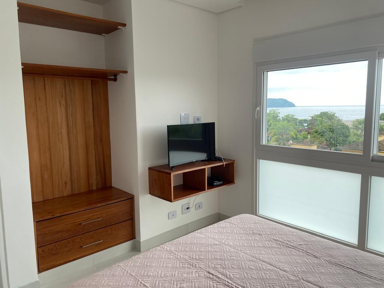 Une chambre à coucher avec un lit, une armoire en bois, un téléviseur posé sur une étagère et une fenêtre donnant sur un beau paysage.