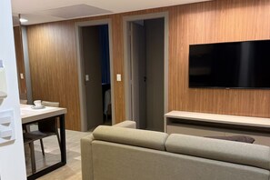 A modern hotel room with a sofa, a TV mounted on the wall, a table with chairs, and wooden paneling on the walls. - Meliá Flat Block B 307 (Brasília)
