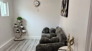 Ein Zimmer mit einem grauen, gepolsterten Sofa, einem kleinen runden Tisch mit goldener Basis, einer Wanduhr und einem Pflanzenregal.