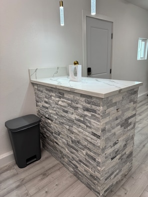 A bathroom with a marble countertop, a stone wall, a black trash bin, and a door in the background.