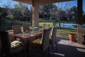 Un patio al aire libre con una mesa de ladrillo y sillas de mimbre, con vistas a un césped y una piscina.