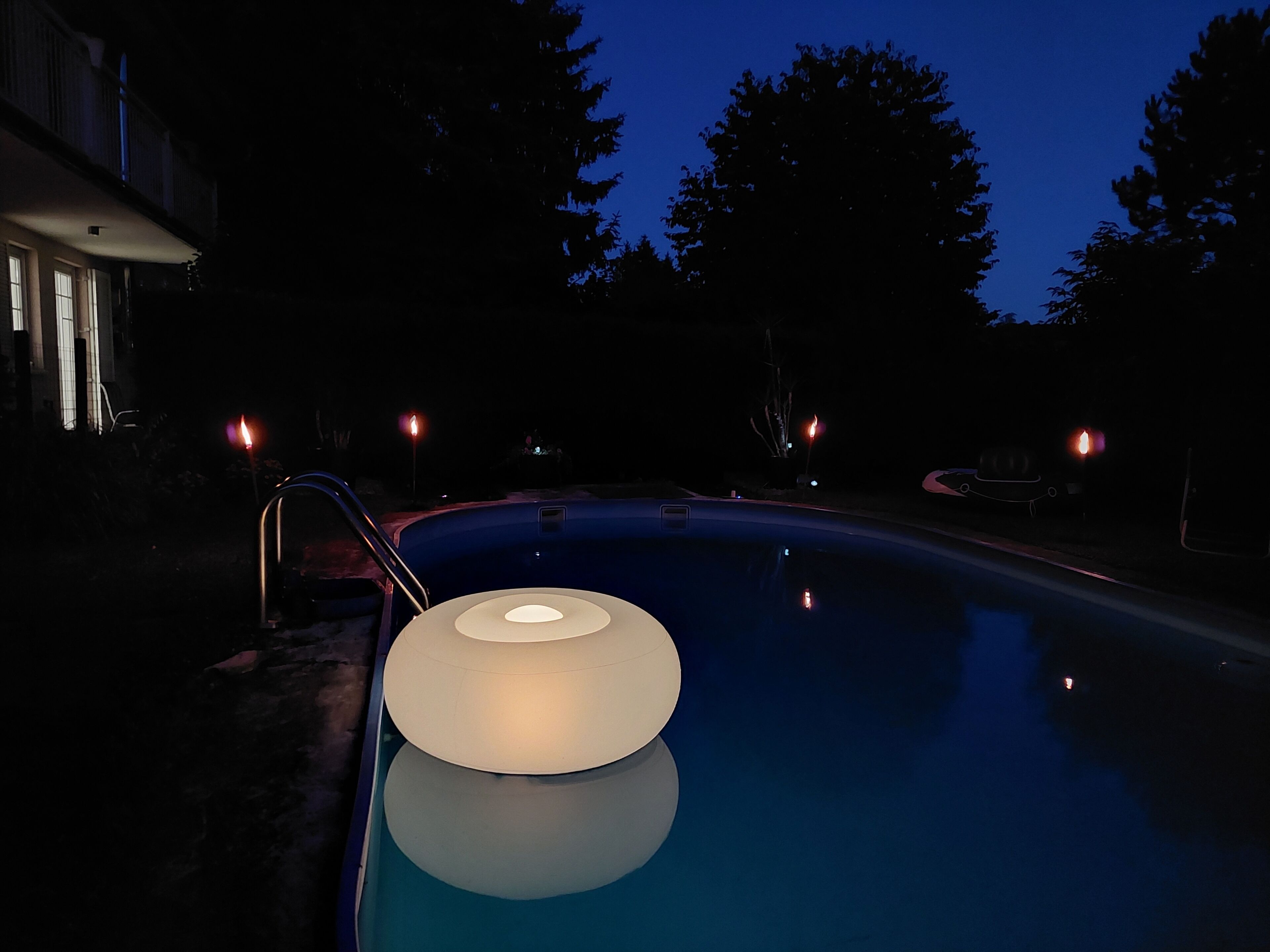 Ein Swimmingpool mit einer runden Leuchte in der Mitte, umgeben von Bäumen und einem Gebäude im Hintergrund.