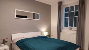 A neatly made bed with a teal bedspread, a bedside table with a lamp, a mirror on the wall, and a window with curtains. - HC 104 big apartement with pool and garden (Haidershofen)