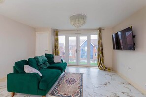 A living room with a green sofa, a patterned rug, a chandelier, and a TV on the wall. - The Meridian Langley (Slough)