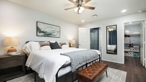 Une chambre à coucher comprenant un lit, des tables de chevet, un banc, un ventilateur de plafond, une douche et un dressing.