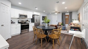 Une cuisine moderne avec des meubles blancs, des appareils en acier inoxydable, un coin repas avec une table ronde et des chaises en bois, et un salon avec un canapé et une lampe.