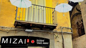 Un restaurante de sushi con varios paraguas afuera, un cartel que indica que ofrece comida para llevar y un tablero rojo con el menú.
