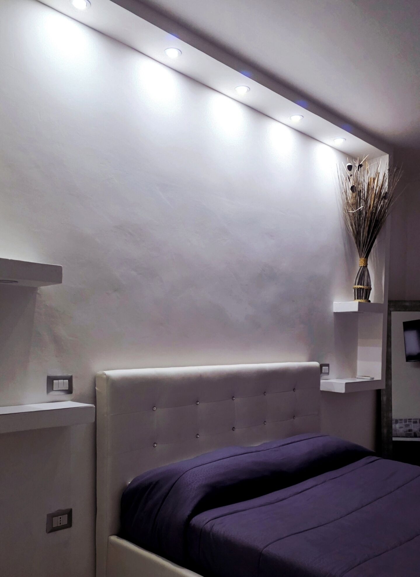 Une chambre moderne avec un lit, des spots encastrés au plafond, une étagère murale ornée d’un vase et une petite étagère sur laquelle repose une télécommande.