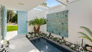 Un área de piscina moderna al aire libre con una puerta geométrica, una pared decorativa y una palmera.