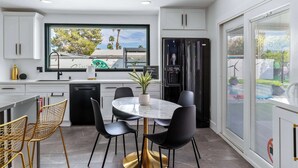 A modern kitchen with white cabinets, a black refrigerator, a dining table with chairs, and a large window. - 4BR Resort Home w Pool, Hot Tub & Fire Pit (Palm Springs)