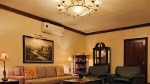 Una sala de estar con sofá, sillones, una mesa de centro, una araña, un cuadro, un aparador y una puerta.