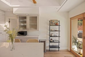 A compact kitchen with a microwave, a white cabinet, a black shelf unit, and a dining area with a table and chairs. - Desert Oasis Home w/ Pool, Pet-Friendly Retreat (Palm Desert)