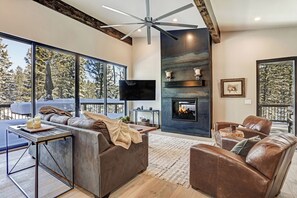 Un salón moderno con chimenea, sofás de cuero, televisor y ventanas amplias con vistas a un paisaje nevado.